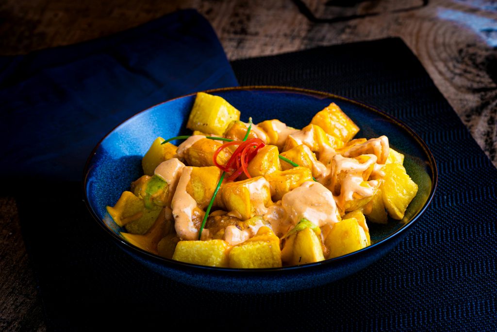Succulent patatas bravas topped with spicy aioli in vibrant blue bowl, served on dark table.